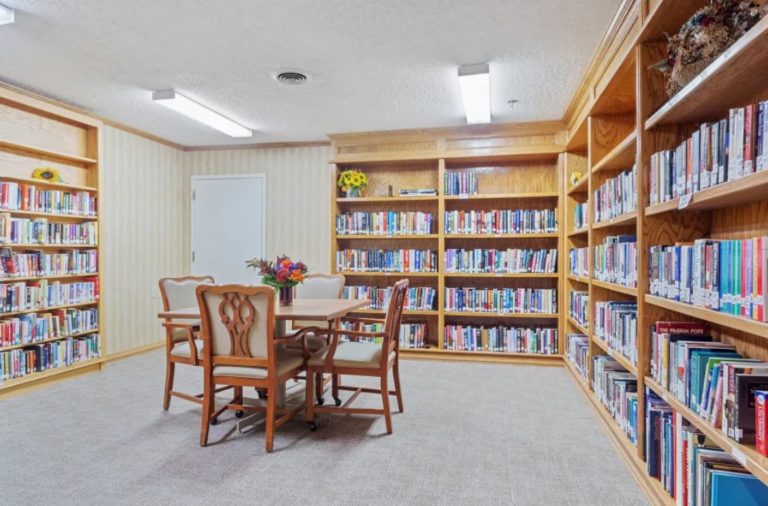 A small library room with wooden shelves filled with books, featuring a table and chairs in the center.