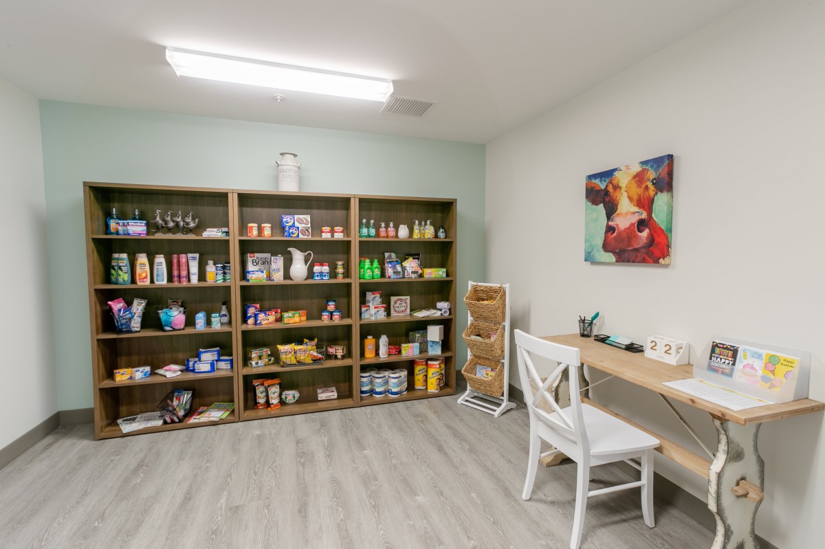 A small store interior featuring shelves stocked with various products, alongside a desk and chair.