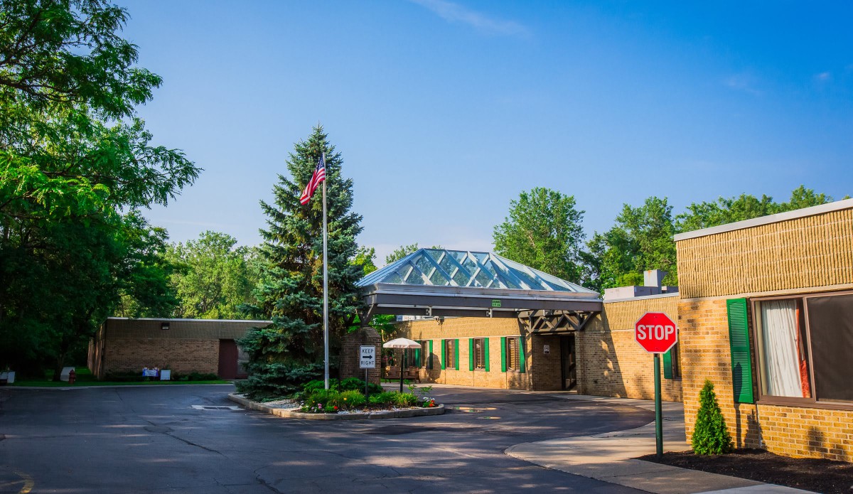 A building entrance with a glass roof, surrounded by trees and featuring a flagpole and a stop sign.