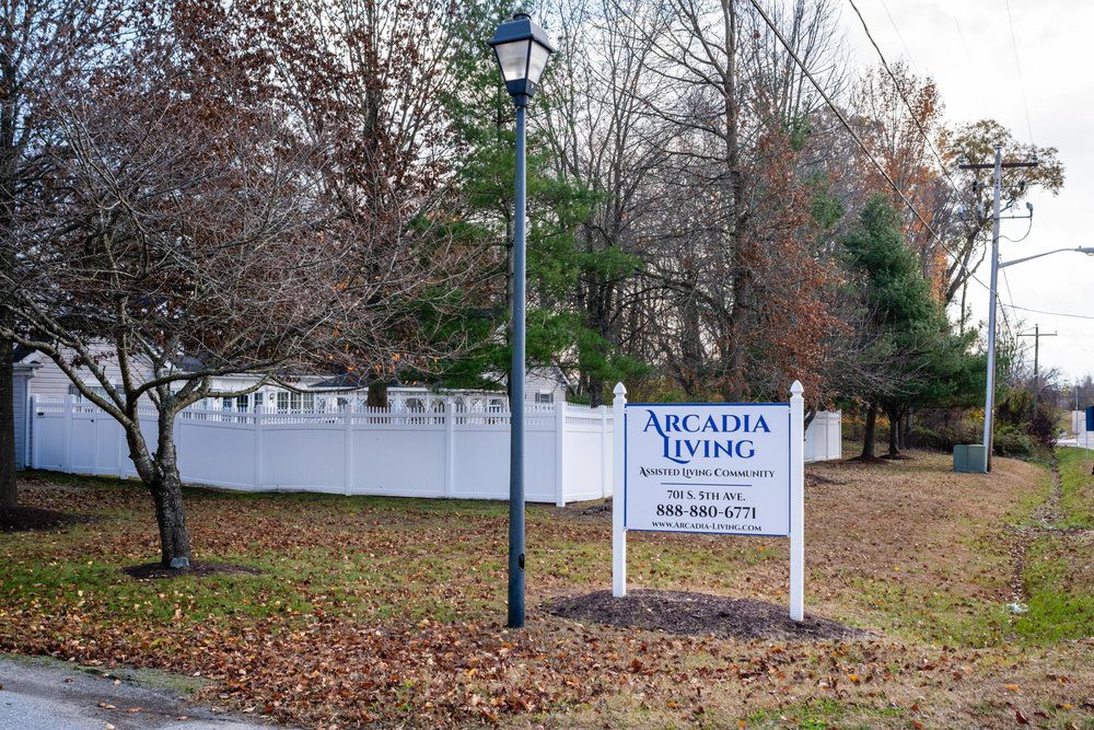 A sign for Arcadia Living stands next to a lamp post, surrounded by a white fence and trees during autumn.