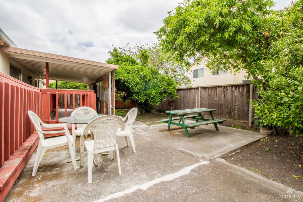 A cozy backyard patio with outdoor seating, including a picnic table and chairs, surrounded by trees and a wooden fence.