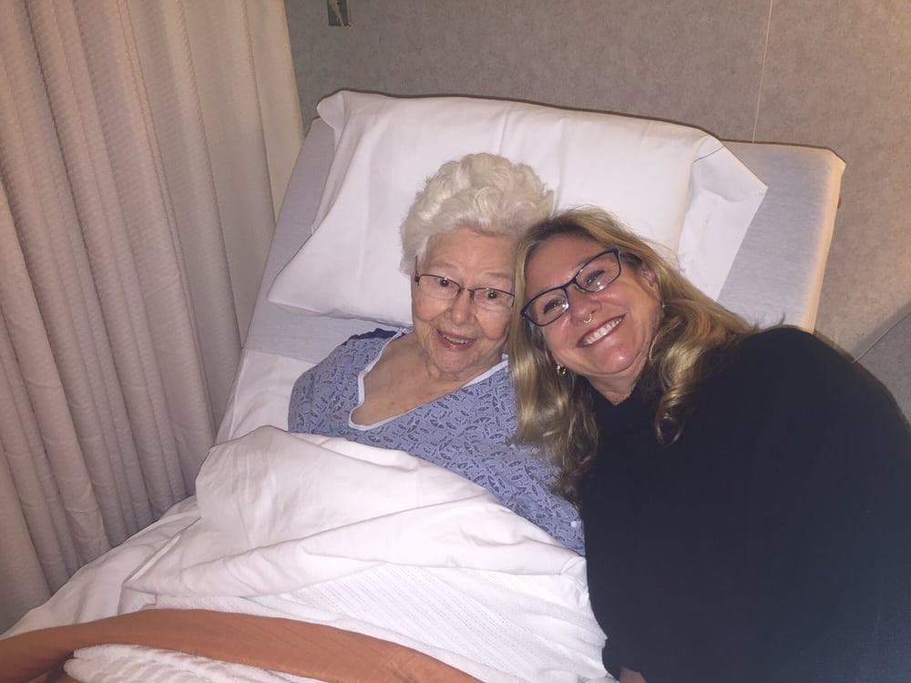 An elderly woman lying in a hospital bed with a younger woman smiling beside her.