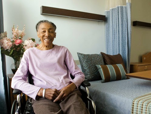 An elderly woman sitting in a wheelchair, smiling warmly in her cozy indoor living space.