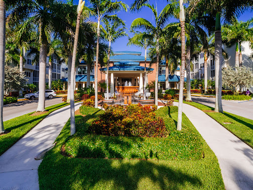A beautifully landscaped entrance to a residential complex, surrounded by palm trees and vibrant flowers under a clear blue sky.