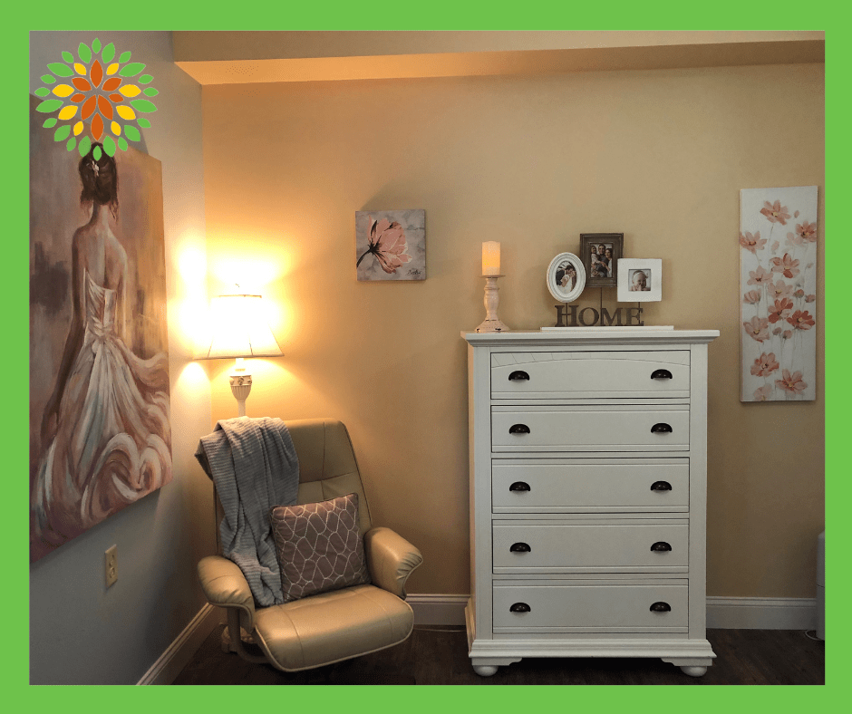 A cozy room with a comfortable chair, a white dresser, and wall art illuminated by a warm lamp.
