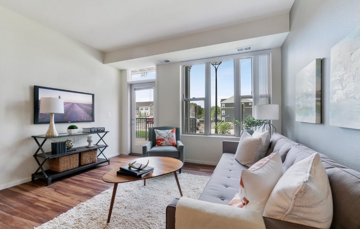 A modern and bright living room with large windows, featuring a comfortable sofa, coffee table, TV stand, and decorative accents.