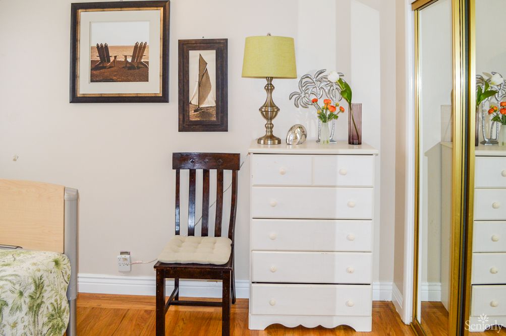 A cozy corner with a white dresser, wooden chair, decorative lamp, and artwork on the wall.