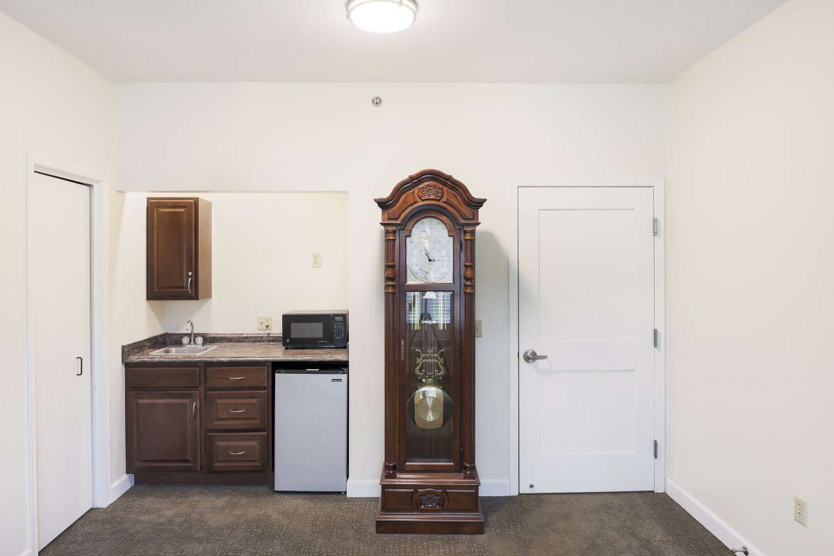 A room interior featuring a grandfather clock next to a kitchenette with a mini fridge and microwave.