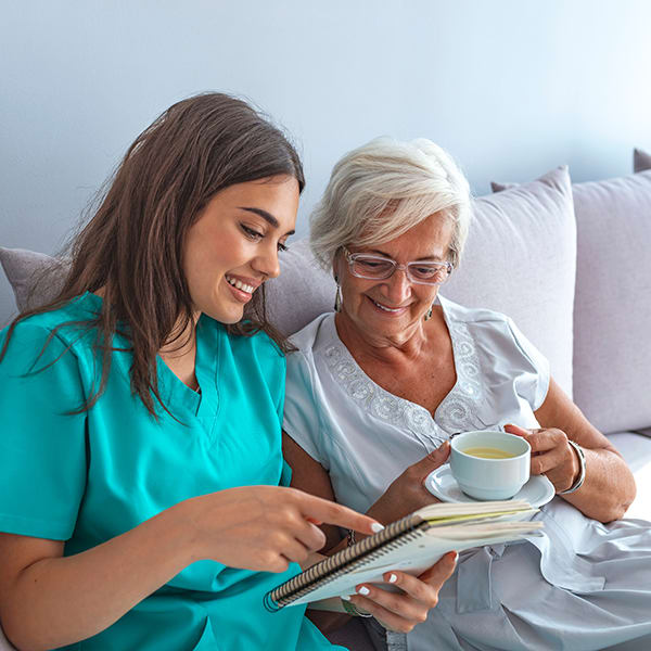 A nurse and an elderly woman enjoying a conversation over tea while looking at a book.