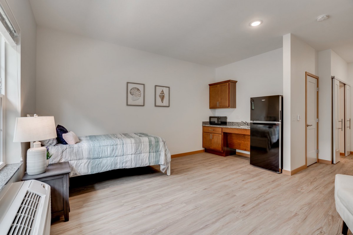 A minimalist bedroom with a single bed and a small kitchenette, featuring a fridge and wooden flooring.