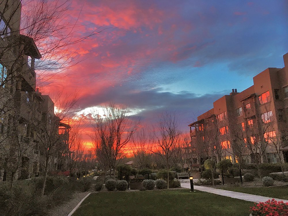 A vibrant sunset paints the sky over apartment buildings, casting a warm glow on the pathway and trees below.