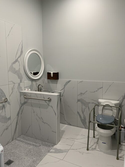 An accessible bathroom with a sink, mirror, toilet, and grab bars, featuring marble tile walls.