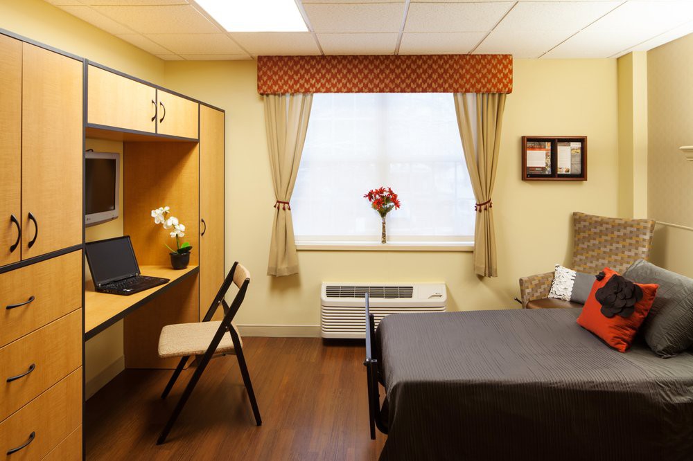 A cozy bedroom with a desk, bed, and window adorned with curtains and flowers.