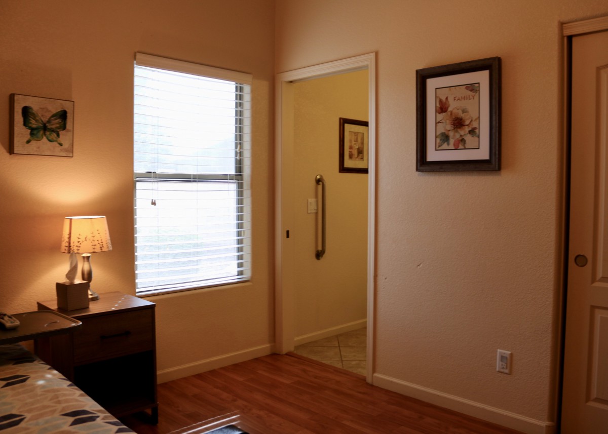 A cozy bedroom with a window, lamp, framed pictures, and a doorway leading to another room.