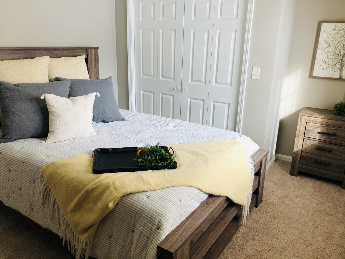 A cozy bedroom with a neatly made bed, featuring a mix of gray and white pillows and a yellow throw blanket, next to a double-door closet.