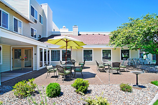A sunny outdoor patio area with tables, chairs, and an umbrella, surrounded by landscaped greenery.