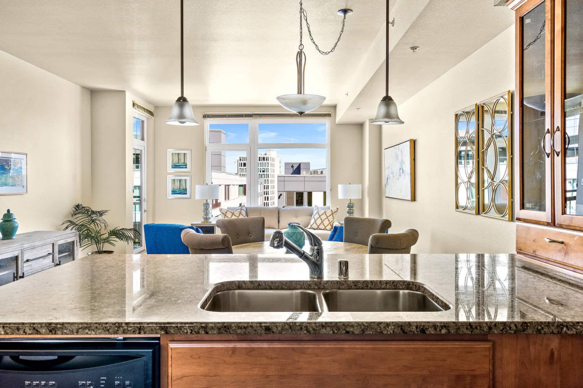 A modern open floor plan featuring a kitchen with a double sink and countertop overlooking a bright living room with large windows.