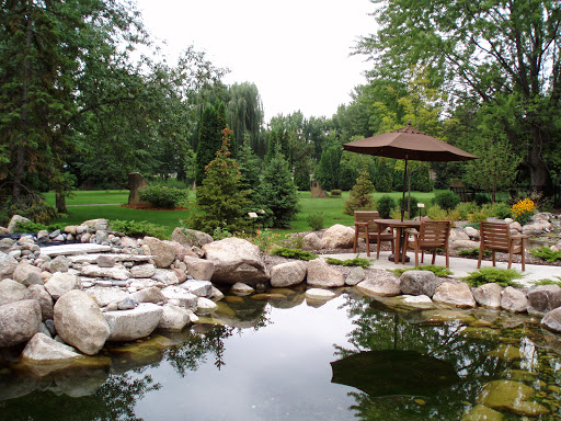 A serene garden scene featuring a tranquil pond surrounded by natural stones and lush greenery, complete with a cozy seating area under an umbrella.