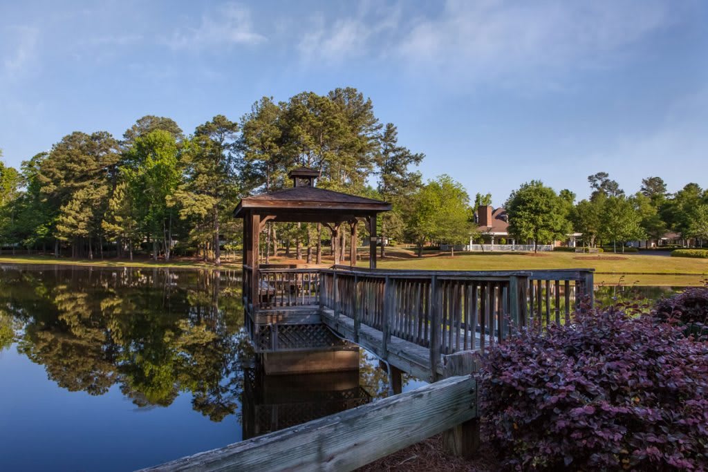 A tranquil scene featuring a gazebo by a pond with a wooden dock extending over the water, surrounded by lush trees.