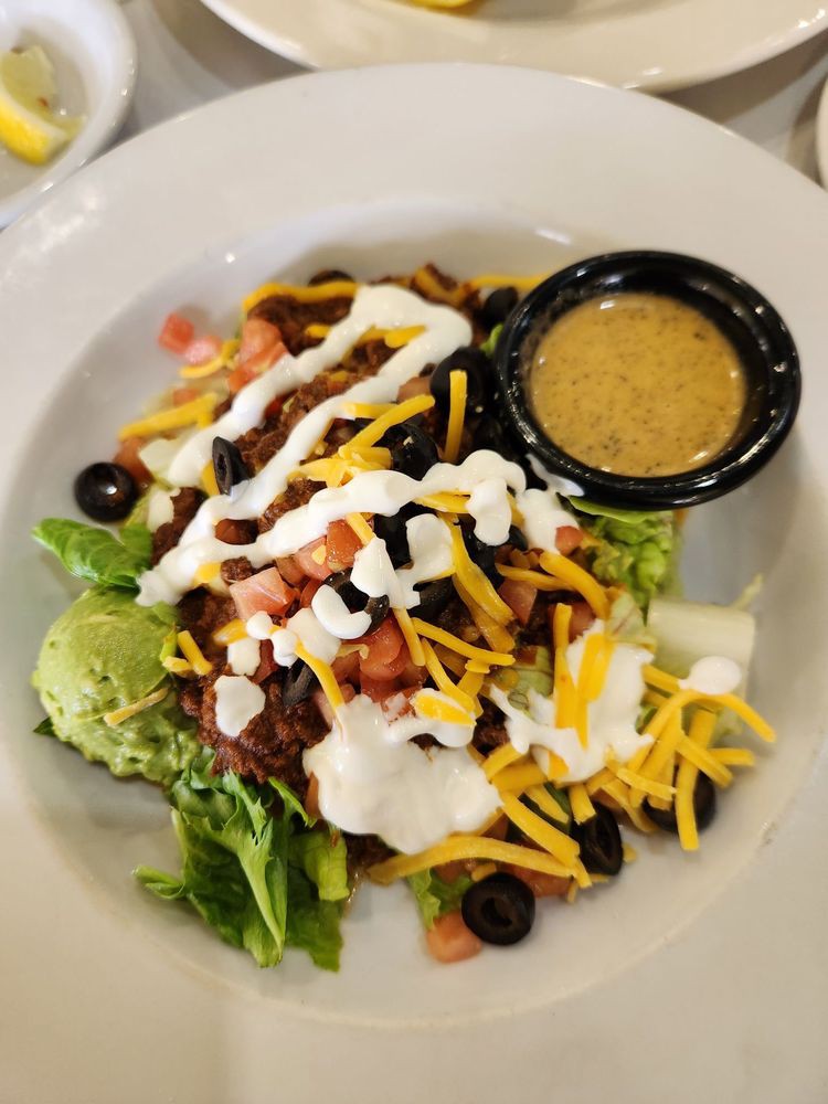 A colorful taco salad topped with cheese, sour cream, and olives, served with a side of dressing.
