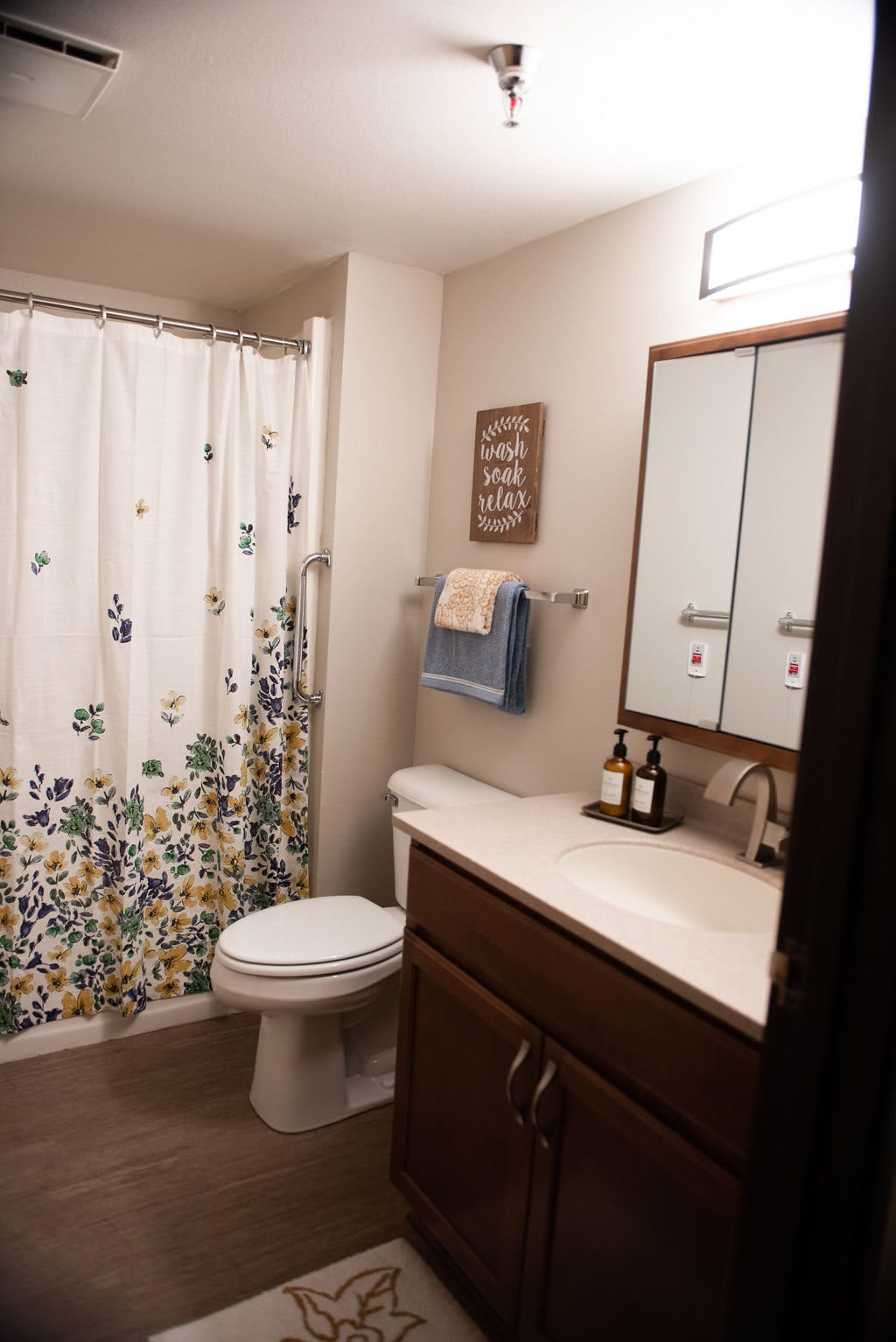 A modern bathroom with a floral shower curtain, white toilet, wooden vanity with a mirror, and neatly arranged towels and toiletries.