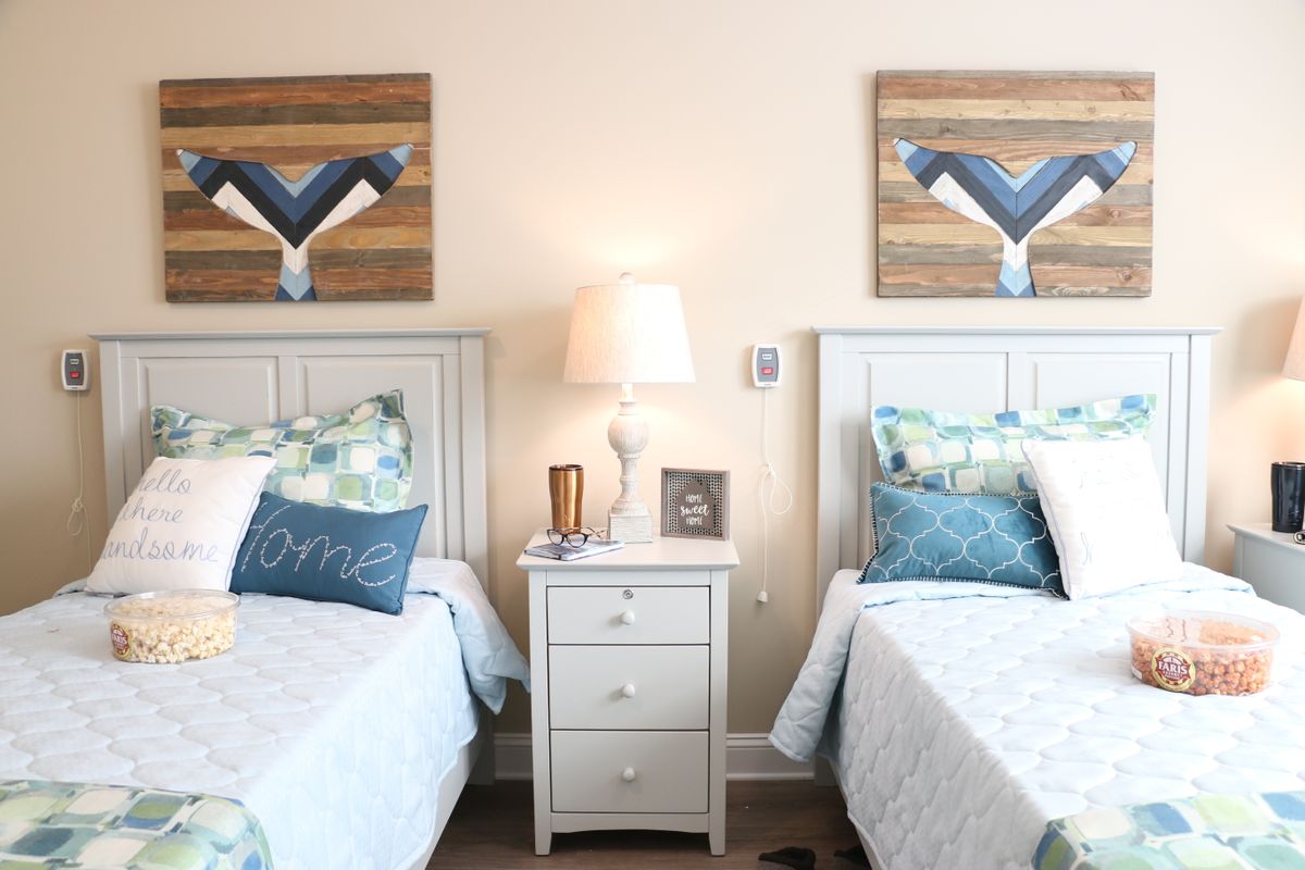 A cozy bedroom with two twin beds featuring blue and white bedding, a central nightstand with a lamp, and decorative artwork above each bed.