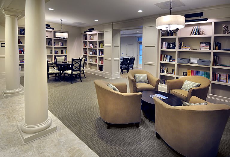 A cozy library with armchairs and bookshelves, featuring warm lighting and a carpeted floor.