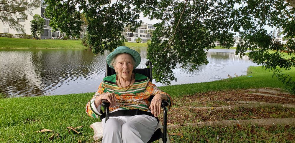 An elderly woman in a wheelchair enjoys a day outdoors by the river, smiling under the shade of trees.
