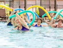 A group of people participating in a water aerobics class, using pool noodles for exercise.