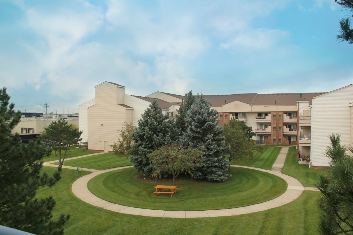 A serene park area with neatly trimmed grass and a circular pathway, surrounded by trees, next to a building under a partly cloudy sky.