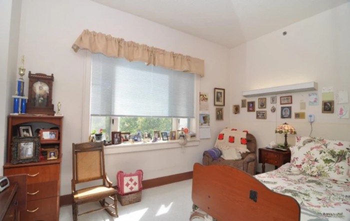 A cozy bedroom featuring a bed, chair, window with curtains, and walls adorned with photos.