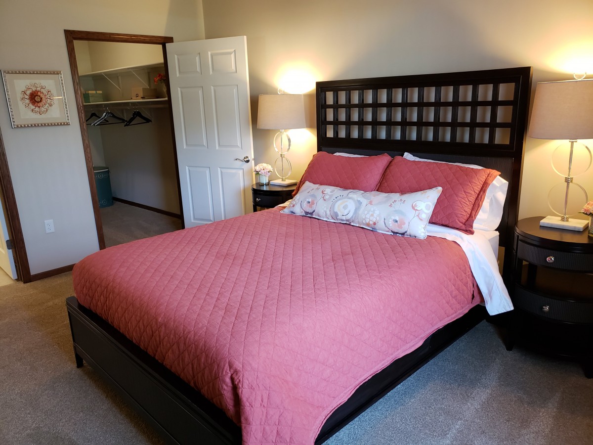 A cozy bedroom with a bed featuring a pink cover and decorative pillows, accompanied by a nightstand with a lamp and an open closet door.