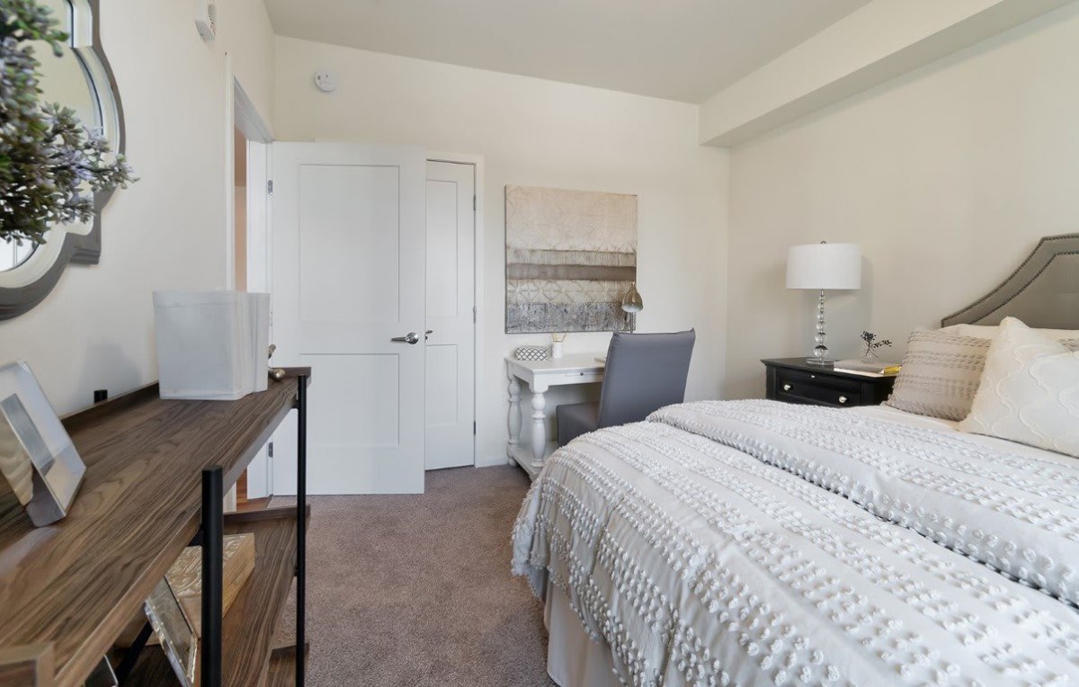 A cozy bedroom featuring a bed with neutral-colored bedding, a desk with a chair, and a lamp on the nightstand.