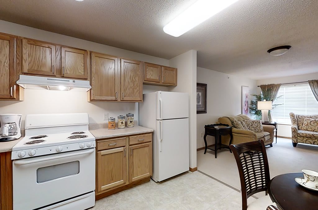 An open-layout kitchen in an apartment, featuring wooden cabinets and essential appliances including a refrigerator and stove.