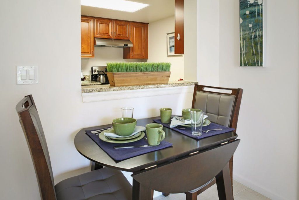 A modern dining area with a wooden table set for two, adjacent to a kitchen with wooden cabinets.