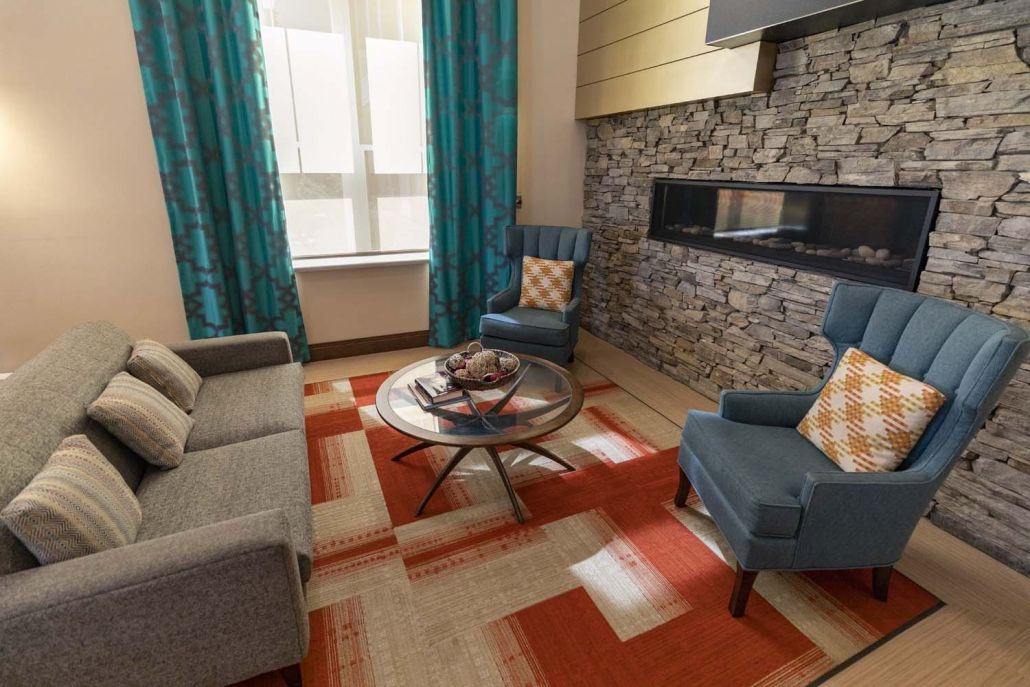 A cozy living room featuring a modern stone fireplace, blue armchairs, and a vibrant orange rug.