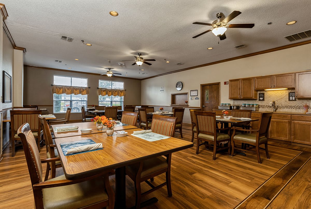 A spacious dining area with wooden flooring, ceiling fans, and a kitchen in the background.