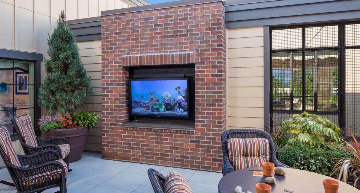 An outdoor patio with a brick wall featuring a mounted television and comfortable seating.