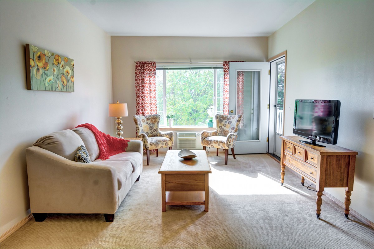 A cozy living room with a sofa, two armchairs, and a coffee table facing a TV, with sunlight streaming through a large window.
