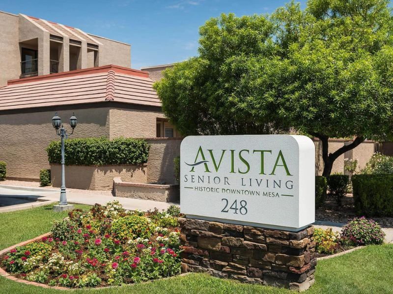 The image displays the entrance sign of Avista Senior Living, set against a backdrop of well-maintained landscaping and residential buildings.