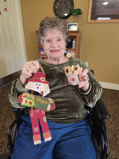 An elderly woman proudly displays her handcrafted scarecrow and decoration, showcasing her creativity and holiday spirit.