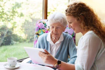 A young woman assists an elderly woman with using a tablet indoors.