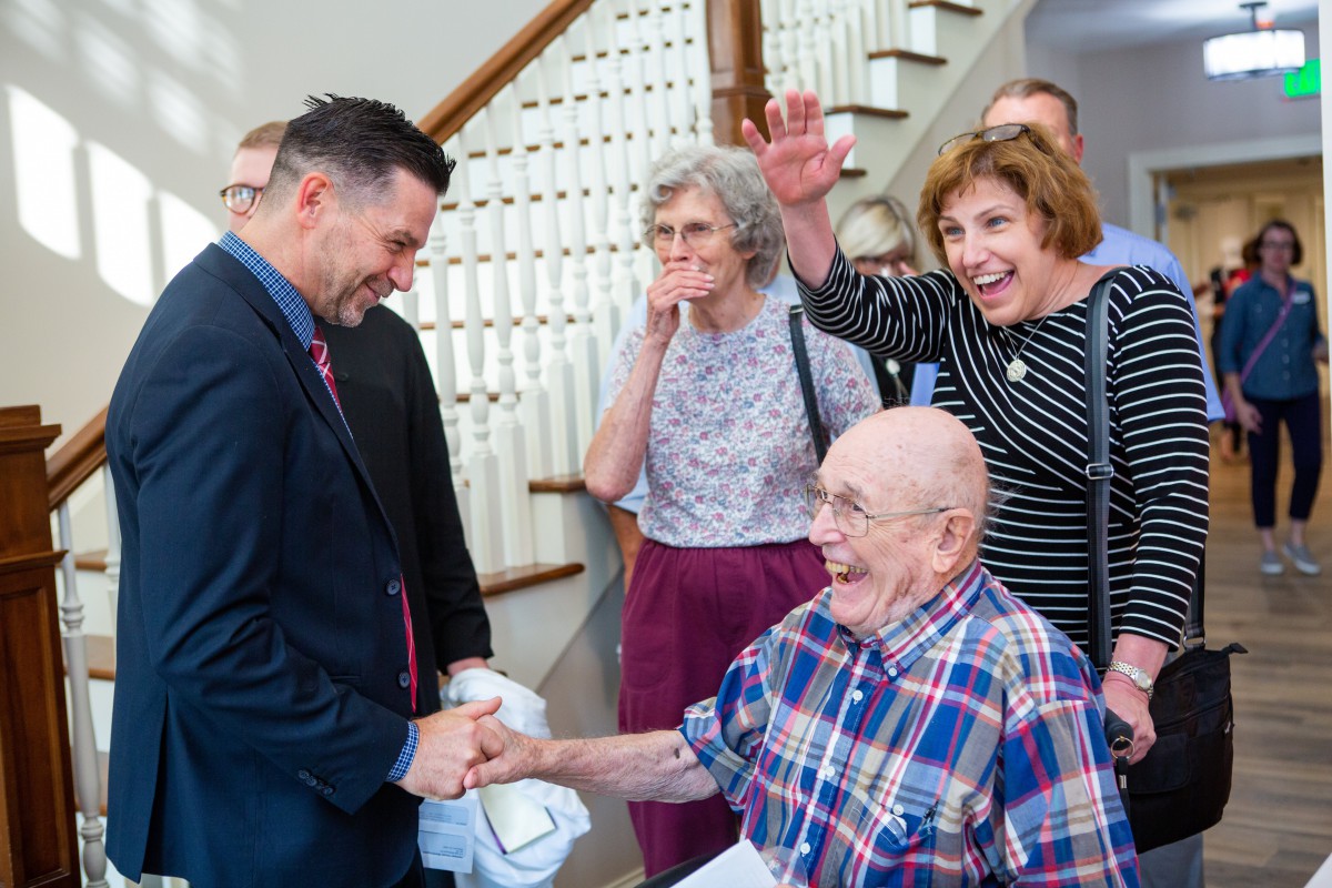 A group of people gather indoors as a man in a suit greets an elderly gentleman with a handshake, surrounded by others smiling and interacting near a staircase.