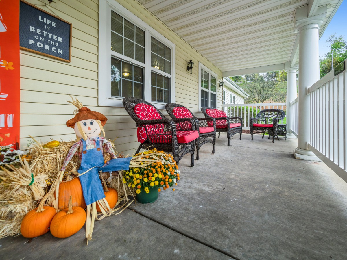 A cozy porch decorated for autumn with a scarecrow, pumpkins, and vibrant flowers.