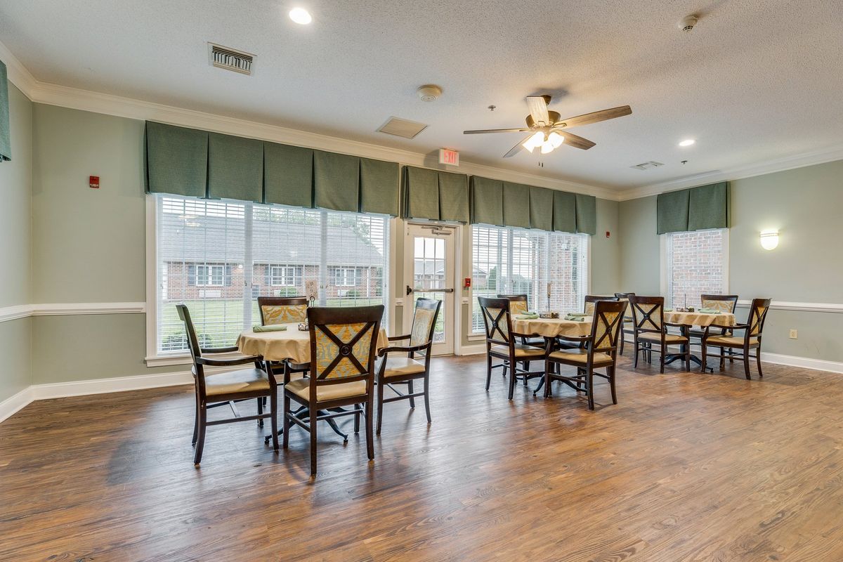 A spacious dining room with wooden flooring, featuring multiple tables and chairs arranged neatly, large windows that allow natural light to flood in, and ceiling fans for ventilation.