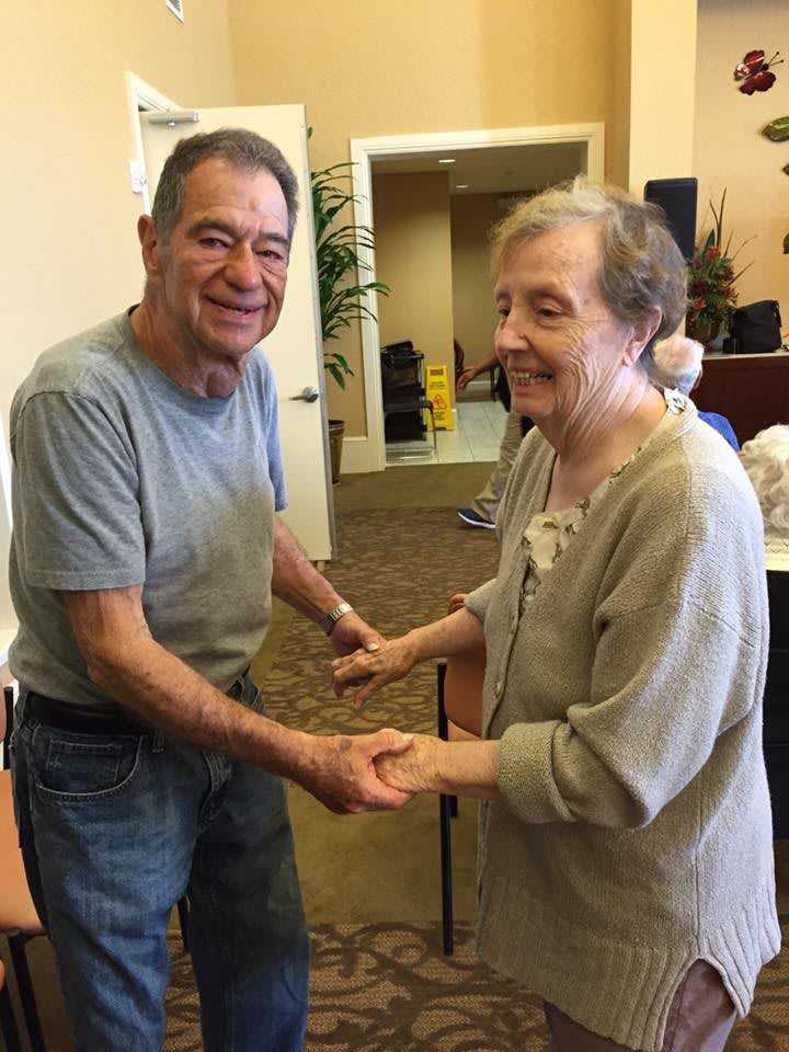 An elderly couple dancing together indoors, holding hands and smiling.