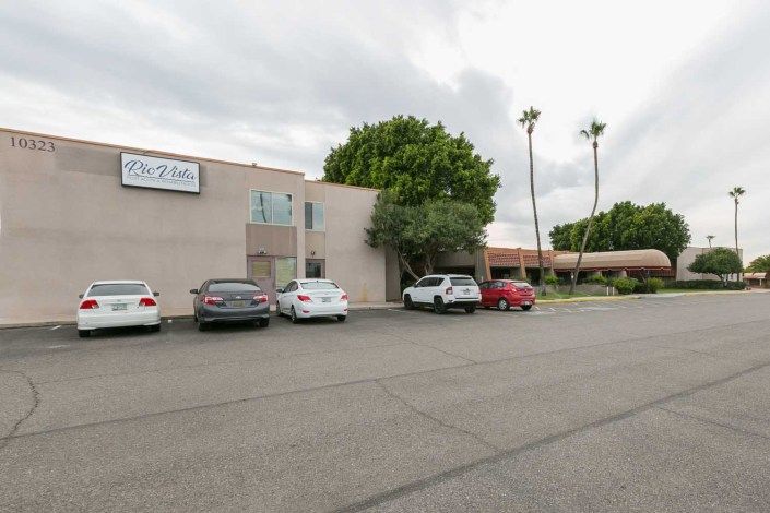A view of a building with a parking lot in front, featuring several cars and palm trees under a cloudy sky.