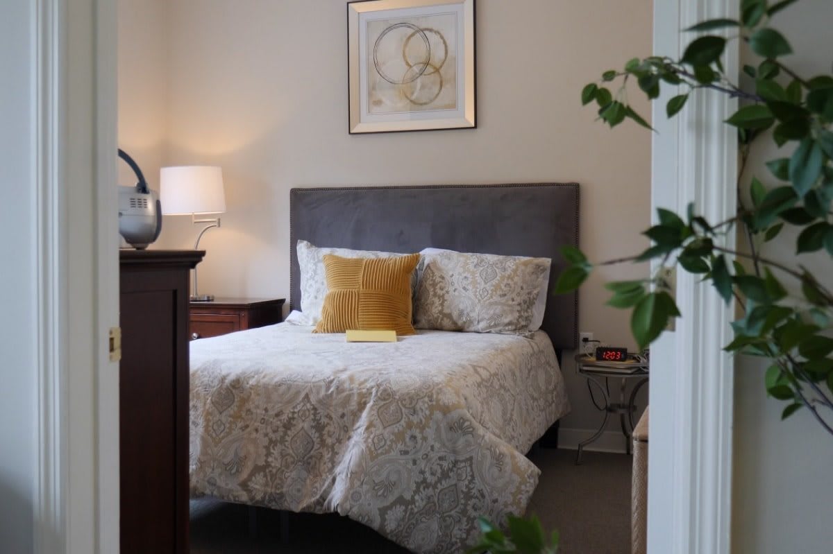 A cozy bedroom featuring a neatly made bed with patterned bedding and decorative pillows, complemented by a lamp and artwork on the wall.
