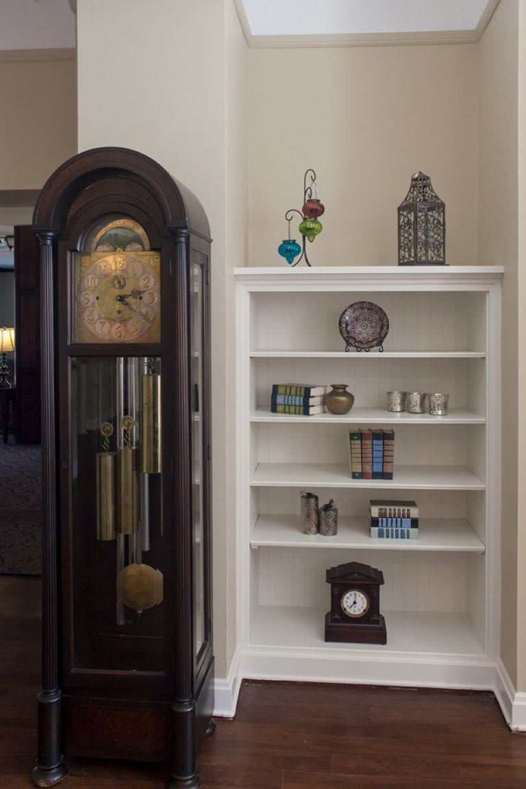 A cozy corner featuring a grandfather clock and a white bookshelf adorned with books and decorative items.