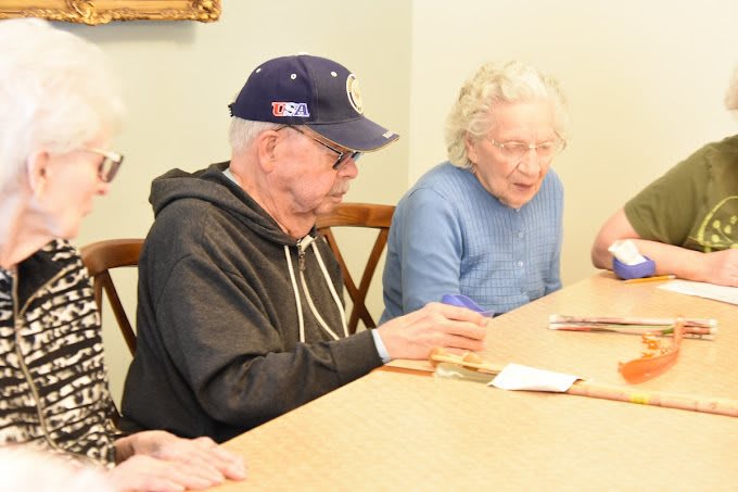 Two elderly individuals are seated at a table, engaged in writing or paperwork.